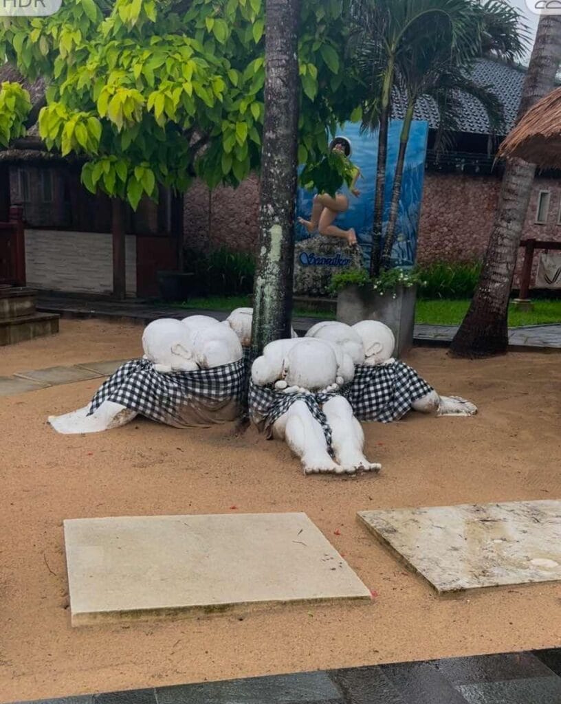 Sculptures of people with bowed heads sitting around a tree trunk evoke a sense of Bali's serene holiness in a tranquil courtyard setting.