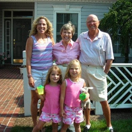 Barb, Grammy Babe and Papa Gordy stand behind Molly and Gracie, smiling together outside a house on a sunny day.